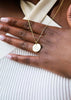 Gold necklace with a round pendant on a hand against a neutral background